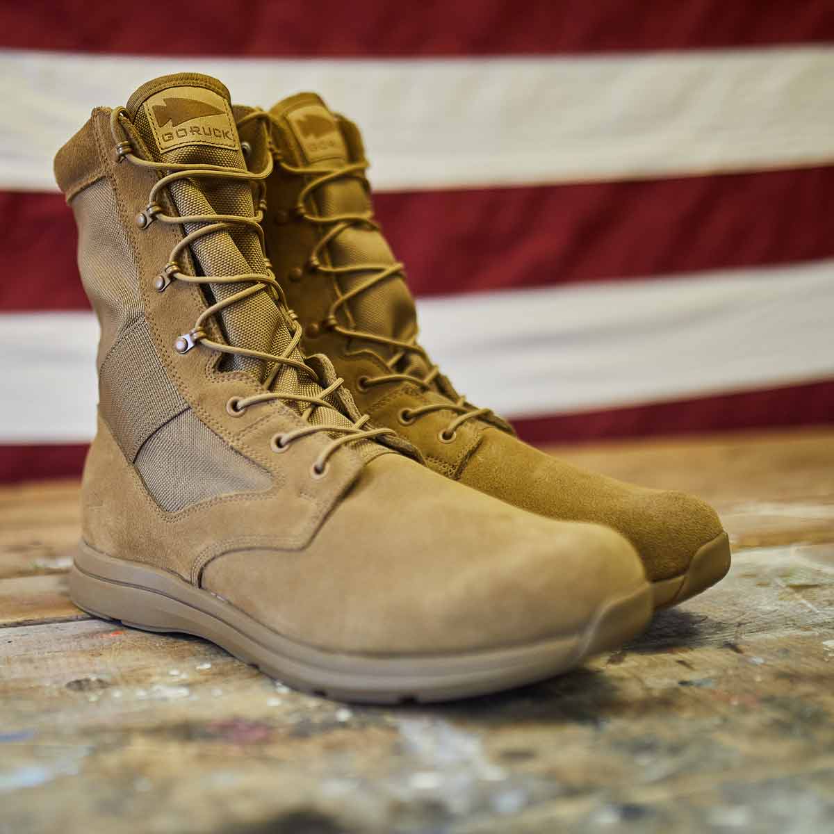 The MACV-1 high-top boots in coyote suede by GORUCK rest on a wooden surface with an American flag blurred in the background. These boots feature an all-terrain outsole and a versatile suede leather upper, with high laces and reinforced seams highlighting their durability.