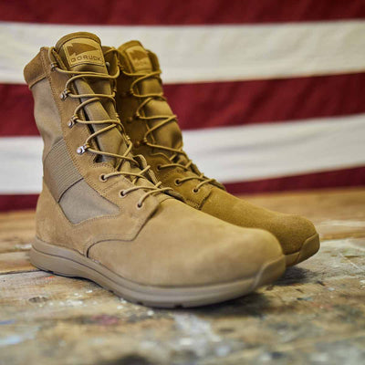 The MACV-1 high-top boots in coyote suede by GORUCK rest on a wooden surface with an American flag blurred in the background. These boots feature an all-terrain outsole and a versatile suede leather upper, with high laces and reinforced seams highlighting their durability.