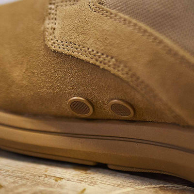 Close-up of the MACV-1 High Top in Coyote Suede by GORUCK, showcasing detailed stitching and a textured suede leather upper. This hiking boot, featuring two round metal eyelets on the side, rests on a wooden surface with its all-terrain outsole designed for optimal performance.