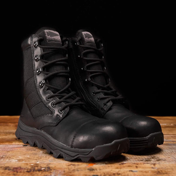 Displayed on a wooden surface against a black background, the MACV-2 Safety Boot - High Top from GORUCK meets Special Forces standards. Designed with laces and side zippers, they exemplify the rugged durability you'd expect from top-tier safety and work boots.