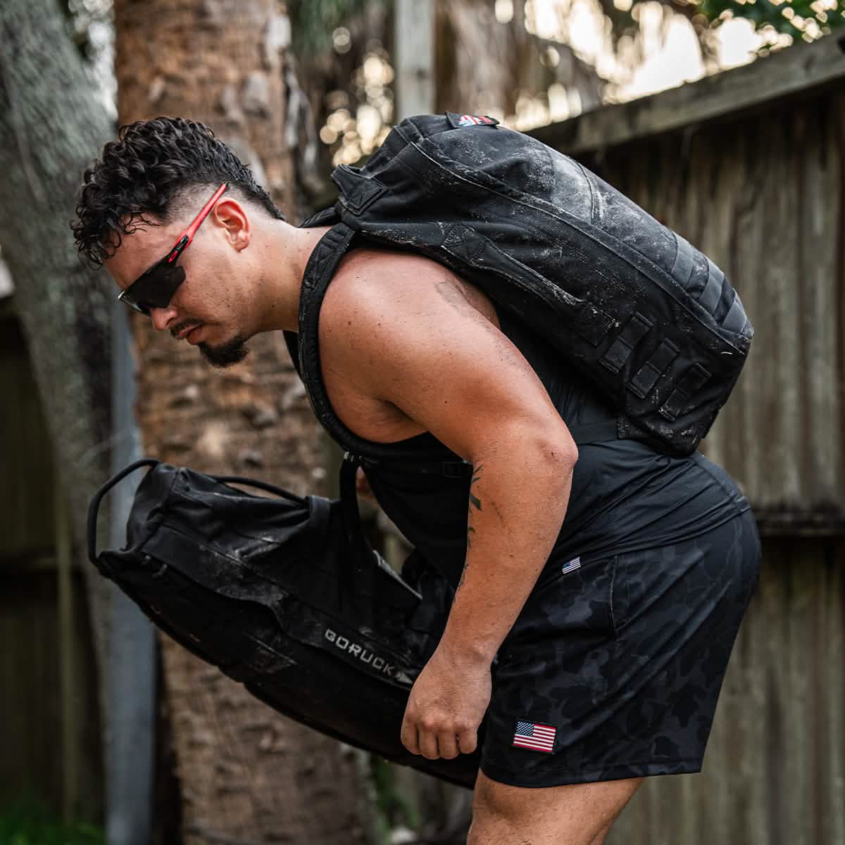 A person wearing a black tank top, shorts, and sunglasses is rucking beside a tree and wooden fence, lifting two dusty GORUCK Ruck Clubs Starter Kit - Light bags. One bag displays the word GORUCK and a small American flag patch, perfectly capturing the adventurous spirit of any GORUCK event.