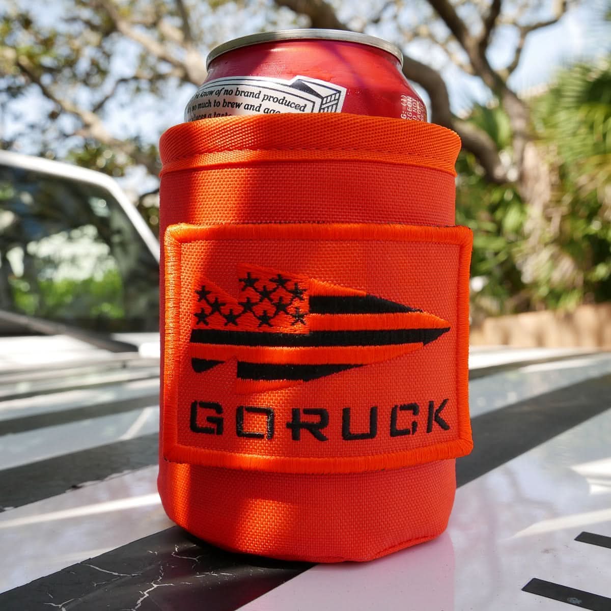 The Ballistic Beer Jacket - GORUCK features black "GORUCK" text and a flag design on an orange koozie made from durable 1000D CORDURA, providing a snug fit around outdoor cans.