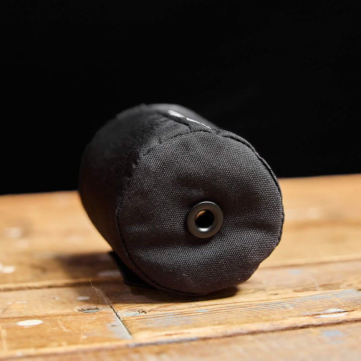 The Ballistic Beer Jacket by GORUCK, a cylindrical black pouch made from durable 1000D CORDURA, includes a metal eyelet and is displayed on a wooden surface against a dark background.