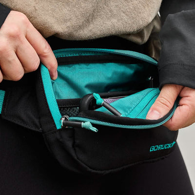 A person is seen wearing a beige top with black sleeves while opening a compact Belt Bag from GORUCK. The black and teal zippered pouch, crafted from durable Cordura fabric, is revealed to contain essentials like a phone and a black carabiner.