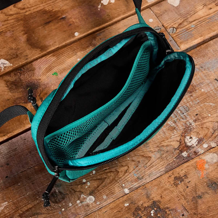 A GORUCK Belt Bag in open green lies on a wooden surface, highlighting its compact design. This crossbody bag includes black zippers, multiple compartments such as mesh pockets, and adjustable webbing for versatility. The wood under the bag displays paint splashes and signs of wear.