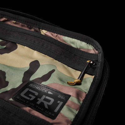 Close-up of a camo-patterned GR1 Birdwell backpack pocket with a black zipper partly open. A small black tag reads GORUCK, highlighting American craftsmanship. A yellow zipper pull complements the design, crafted with durable SurfNyl™ fabric for lasting wear.