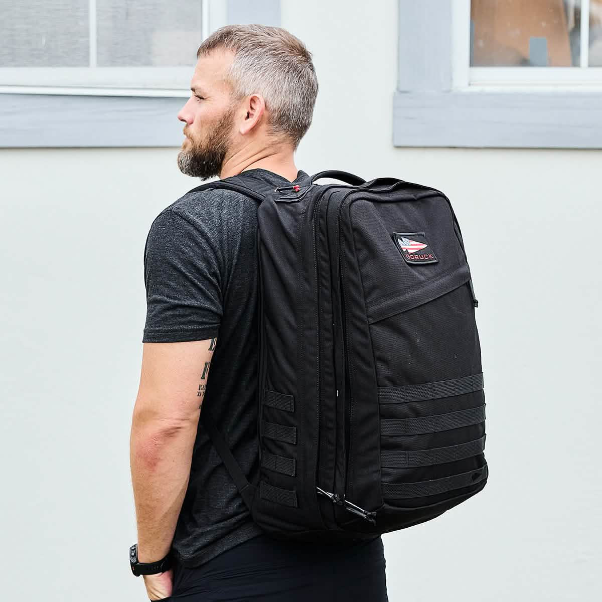 A man with short hair and a beard stands outdoors wearing a black shirt and a large, GORUCK GR2 backpack, known for its Special Forces quality. The carry-on compliant bag showcases a patch and multiple straps. He is posed against a light grey wall with a window above him.