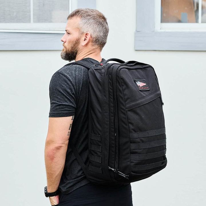 A man with short hair and a beard stands outdoors wearing a black shirt and a large, GORUCK GR2 backpack, known for its Special Forces quality. The carry-on compliant bag showcases a patch and multiple straps. He is posed against a light grey wall with a window above him.