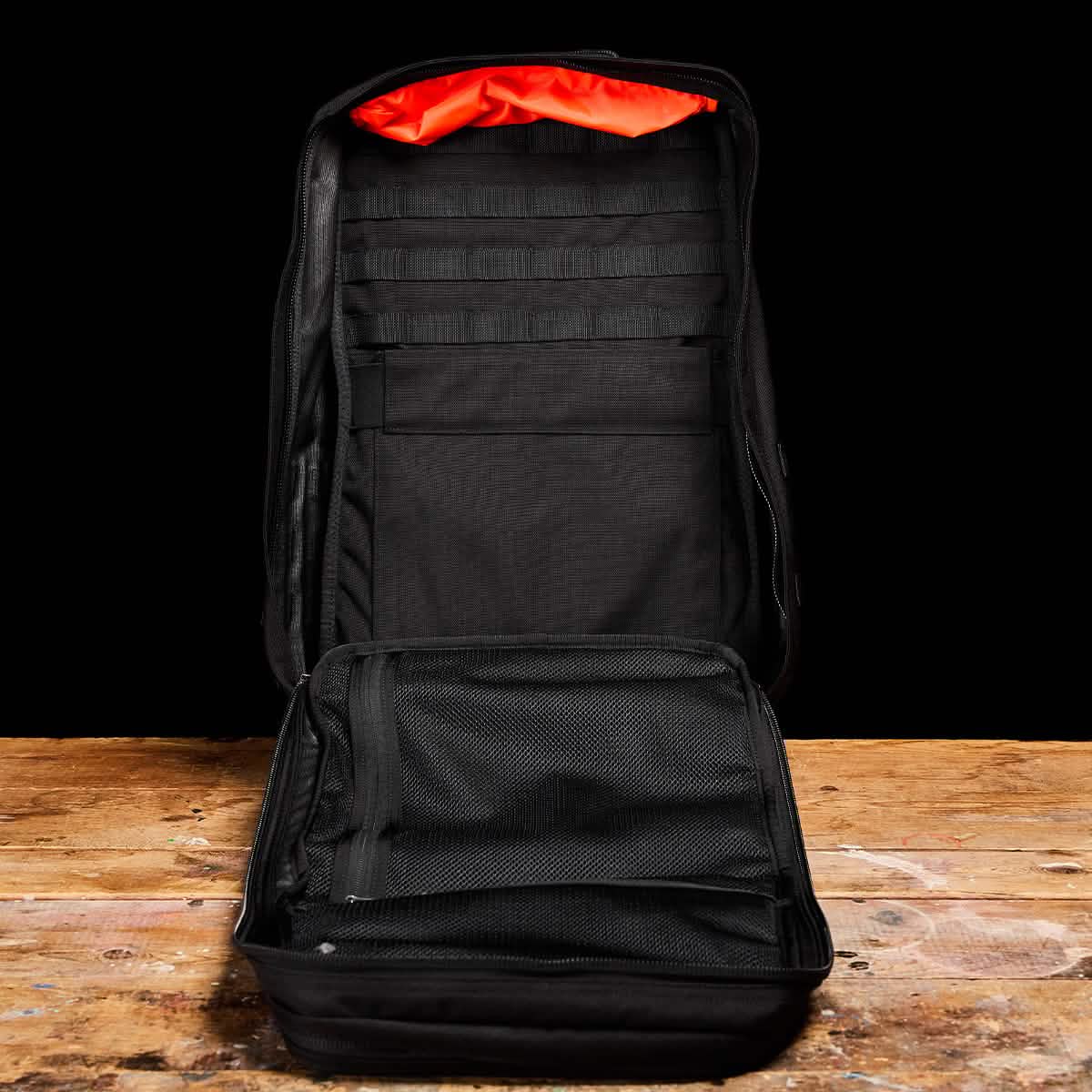 A black GR2 tactical backpack from GORUCK is displayed open on a wooden surface against a dark background. It reveals multiple interior compartments, mesh pockets, and vibrant orange lining - perfect for Special Forces training or everyday ruck adventures.