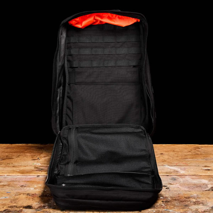 A black GR2 tactical backpack from GORUCK is displayed open on a wooden surface against a dark background. It reveals multiple interior compartments, mesh pockets, and vibrant orange lining - perfect for Special Forces training or everyday ruck adventures.