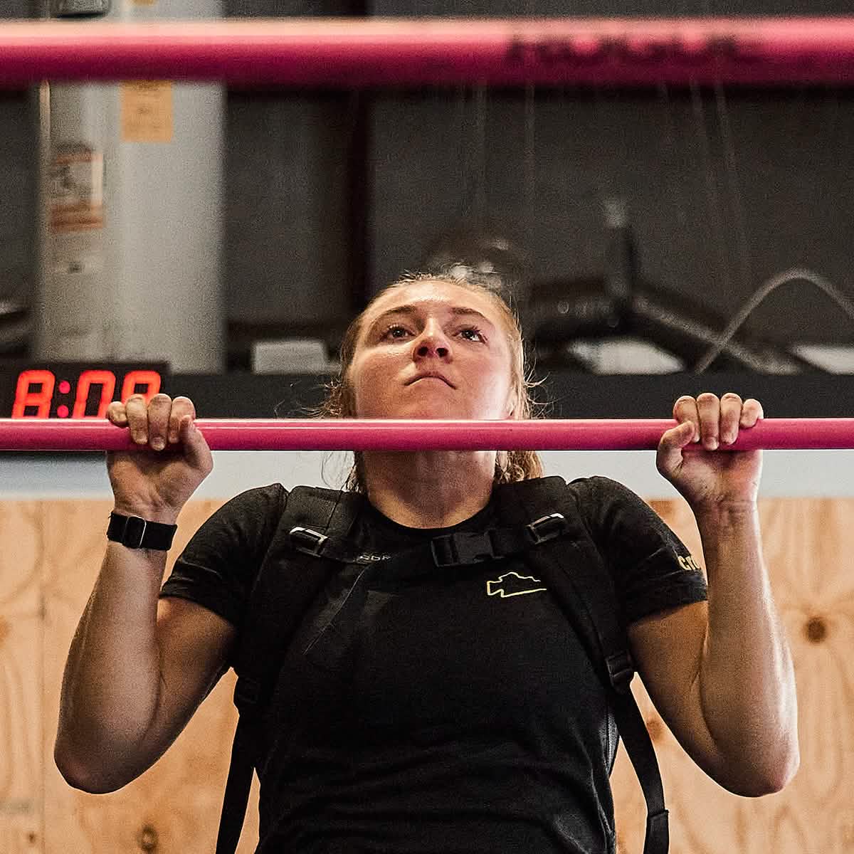 Wearing a GORUCK Women's Subtle Spearhead x CrossFit Cropped Tee in premium poly-blend, paired with a weight vest, an individual executes pull-ups on a pink bar. The backdrop comprises a wooden wall and industrial features, with the red digital clock displaying 8:00 to emphasize the intensity of this workout session.
