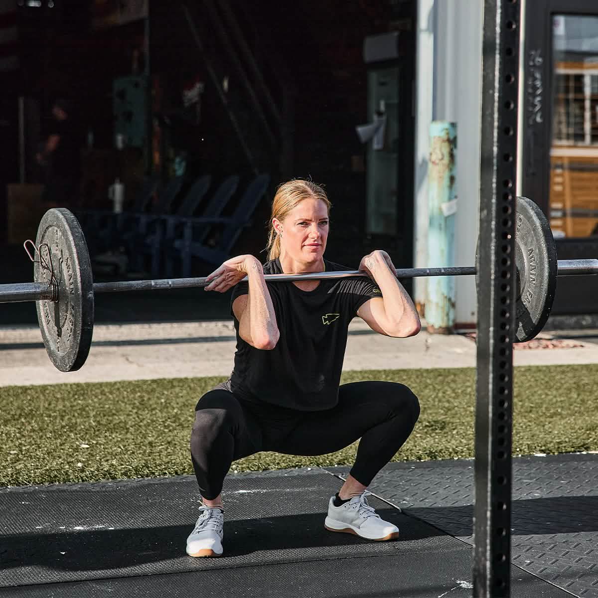 A person confidently performs a front squat with a barbell in an outdoor gym, wearing the stylish Women's Subtle Spearhead x CrossFit Cropped Tee - Poly-Blend by GORUCK and leggings paired with white sneakers. The background reveals workout equipment and a grassy area bathed in bright sunlight, highlighting their dedication.