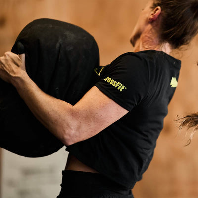 A person lifting a large black sandbag during a CrossFit workout in the Women's Subtle Spearhead x CrossFit Cropped Tee by GORUCK. The gym setting fades into the background, putting full focus on the dynamic action.