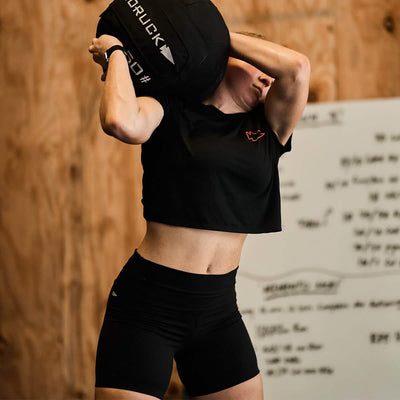 Wearing the GORUCK Women's Subtle Spearhead x CrossFit Cropped Tee in a premium poly-blend, a person lifts a large, black medicine ball over their shoulder in the gym. A wooden wall and a whiteboard filled with writing provide the backdrop to this intense training session.