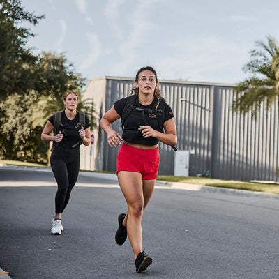 Two women jog down a paved street on a sunny day, displaying their athletic prowess in GORUCK's premium poly-blend clothing and weighted vests. Their workout outfits feature the stylish Women's Subtle Spearhead x CrossFit Cropped Tee by GORUCK. Trees and a large building provide the perfect backdrop for their invigorating run.