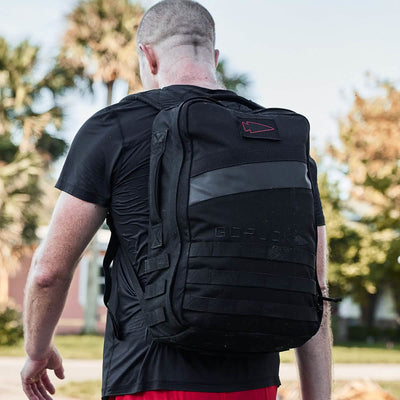 A person with short hair, dressed in a black T-shirt and red shorts, is walking outdoors while rucking. They are carrying a large black Rucker 4.0 rucksack from GORUCK. In the background, there are palm trees and blurred buildings.