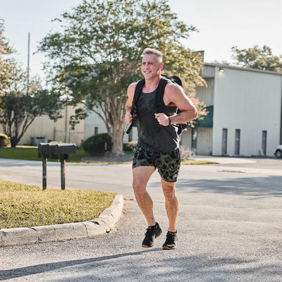A man is jogging on a sunny day, sporting a dark tank top, camouflage shorts, and sneakers. With his Rucker 4.0 backpack by GORUCK strapped securely, he effortlessly combines jogging and rucking. In the background, there's a white industrial building and trees.