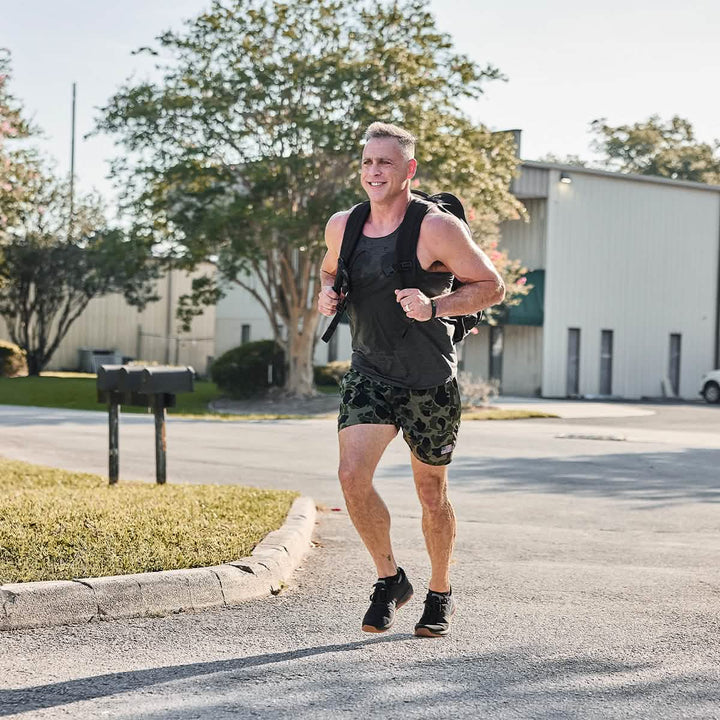 A man is jogging on a sunny day, sporting a dark tank top, camouflage shorts, and sneakers. With his Rucker 4.0 backpack by GORUCK strapped securely, he effortlessly combines jogging and rucking. In the background, there's a white industrial building and trees.