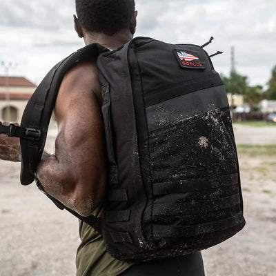 A person with a muscular build carries a GORUCK Rucker 4.0 coated in sand, with its logo prominently displayed. Wearing a sleeveless top, they stand in an overcast outdoor setting with lush greenery and buildings.