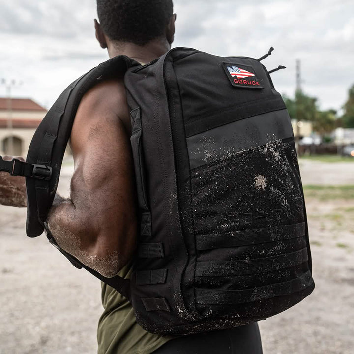 A person with a muscular build carries a GORUCK Rucker 4.0 coated in sand, with its logo prominently displayed. Wearing a sleeveless top, they stand in an overcast outdoor setting with lush greenery and buildings.