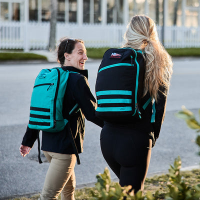 Two people walk outdoors with backpacks, smiling and talking near a white fence and lush greenery. Their Bullet Ruck Classic daypacks, crafted from robust Cordura, add a sense of adventure to the scene.
