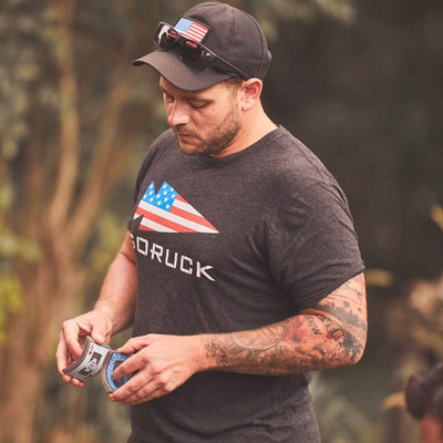 A person wearing a black cap with sunglasses perched on top and a vendor-unknown GORUCK USA Tee - Tri-Blend, featuring an American flag design, examines an energy drink can. With tattoos on their right arm, they stand outdoors in a natural setting, embodying the rugged spirit of GORUCK spearhead adventures.