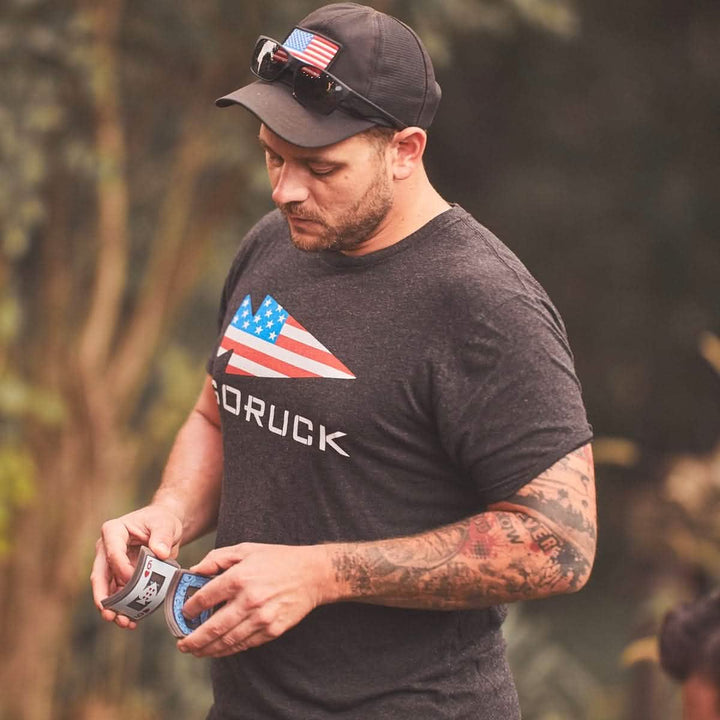 A person wearing a black cap with sunglasses perched on top and a vendor-unknown GORUCK USA Tee - Tri-Blend, featuring an American flag design, examines an energy drink can. With tattoos on their right arm, they stand outdoors in a natural setting, embodying the rugged spirit of GORUCK spearhead adventures.