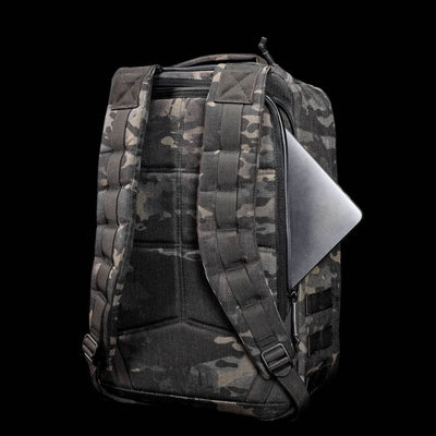 A camo-patterned GR1 USA - Cordura (The Original Ruck) backpack, made with Special Forces-grade materials, features a laptop partially visible in the side zipper pocket, set against a black background.