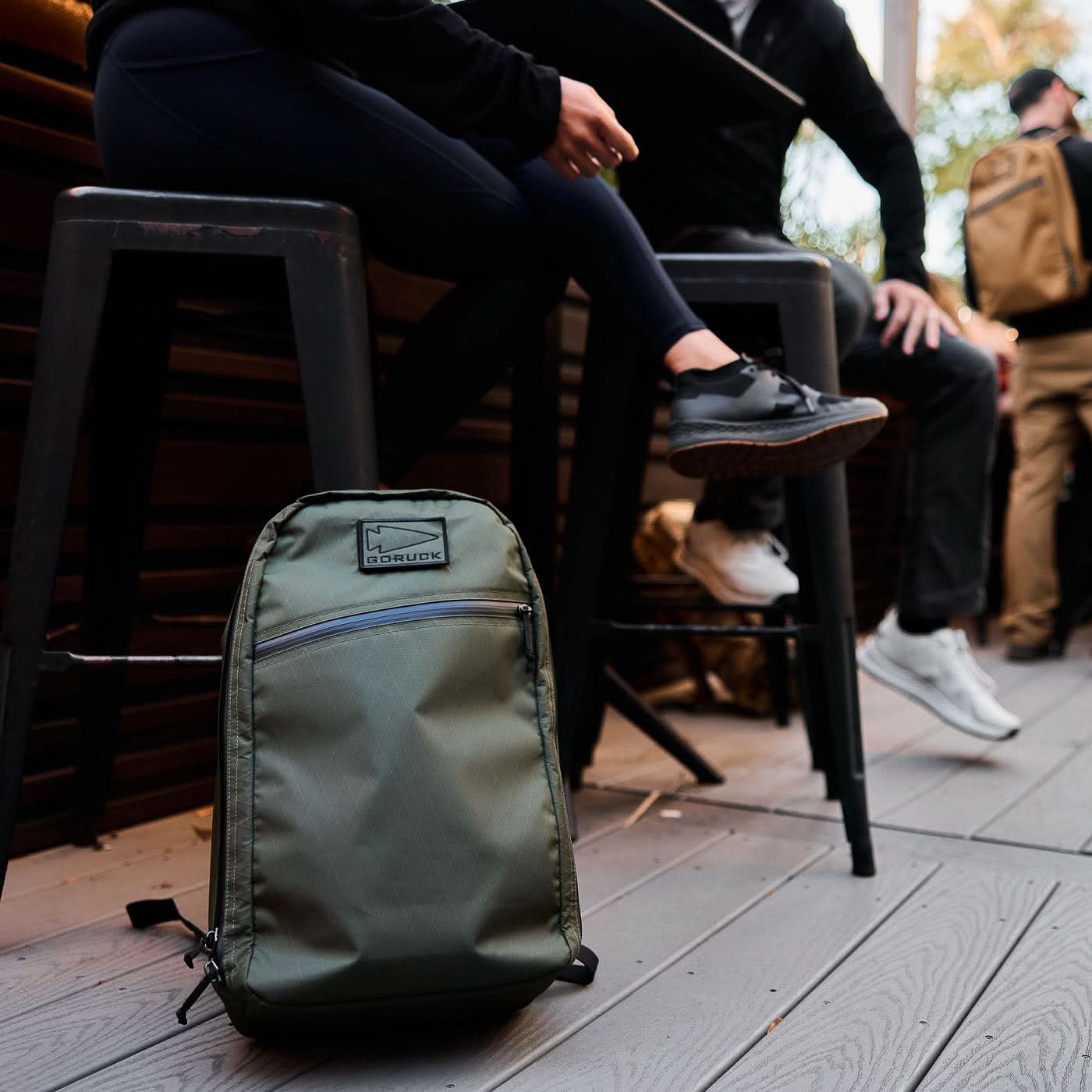 A Bullet X-PAC daypack in green rests on the floor beside two people seated on high stools at an outdoor cafe.