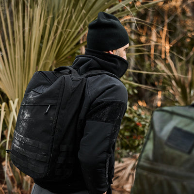 A person in a Men's Task Force Dagger Jacket - Polartec Fleece, wearing a black beanie and carrying a black backpack, stands outdoors near lush foliage, evoking Special Forces resilience.