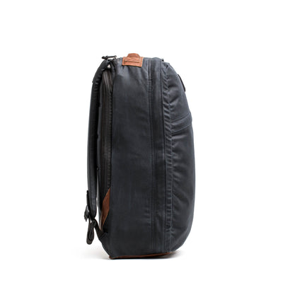 The side view showcases the GR1 USA - Heritage backpack by GORUCK, exhibiting a sleek, dark gray design with leather accents and a minimalist aesthetic made from waxed duck canvas. It includes multiple zippers, a top handle, and adjustable shoulder straps. The backpack is photographed against a plain white background.
