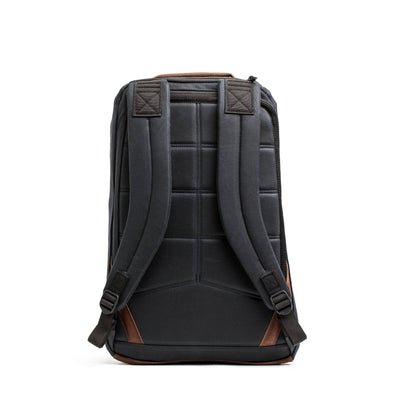 A front view of the GR1 USA - Heritage rucksack by GORUCK, showcasing a black design with brown accents, including padded straps and a quilted back panel. Made from waxed duck canvas, it stands upright against a white background, offering a blend of retro charm and modern functionality.