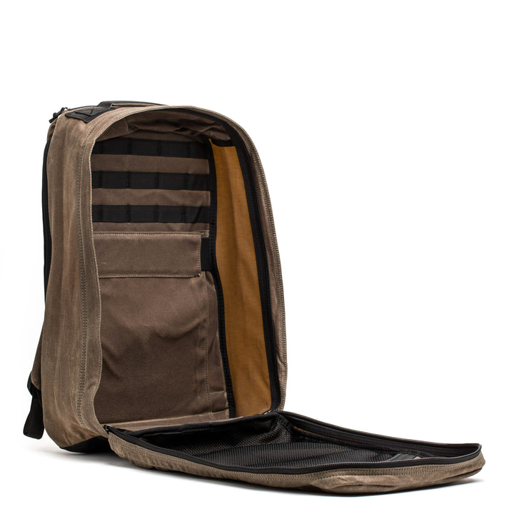 The GR1 USA - Heritage by GORUCK is a brown rucksack with a fully open front zipper that reveals multiple compartments and pockets. Its interior boasts black straps for securing items. Crafted from waxed duck canvas, this rucksack stands upright against a plain white background, exuding vintage charm.