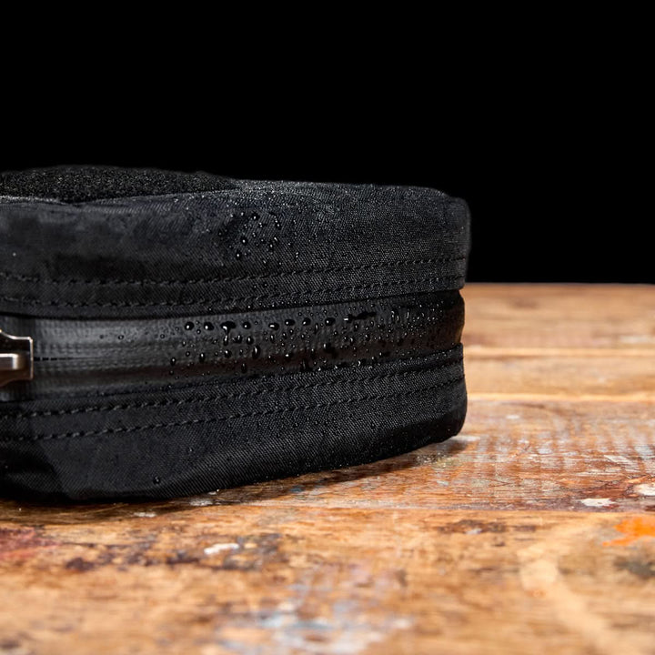 Close-up of the black GR1 Field Pocket - X-PAC electronics organizer, made from waterproof X-PAC fabric, showing water droplets on its AquaGuard zippers while resting on a rustic wooden surface.
