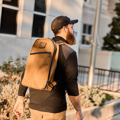 Man with a brown Bullet X-PAC daypack and black cap walking outdoors near a building in sunlight.