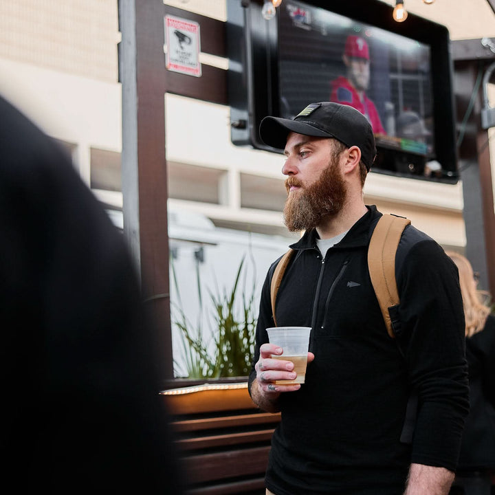 A man with a beard carrying the Bullet X-PAC daypack holds a drink outdoors, while a TV in the background displays sports.
