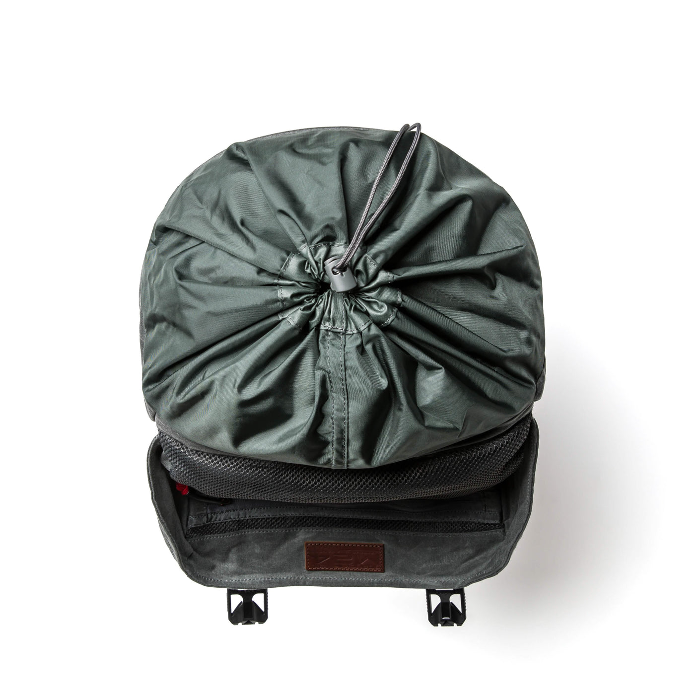 A green M24 rucksack from GORUCK, featuring a drawstring closure on top, is open to display its spacious interior. Made from robust waxed canvas, two small straps dangle from the front flap, and it is set against a plain white background for the photograph.