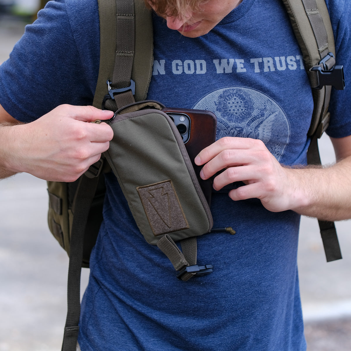 A man in a blue "In God We Trust" shirt puts his phone into the Sternum Pouch - Cordura, attached to his backpack strap, highlighting this handy rucking accessory.