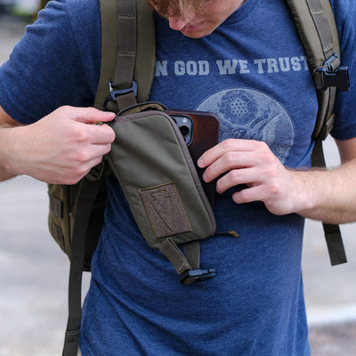 A man in a blue "In God We Trust" shirt puts his phone into the Sternum Pouch - Cordura, attached to his backpack strap, highlighting this handy rucking accessory.