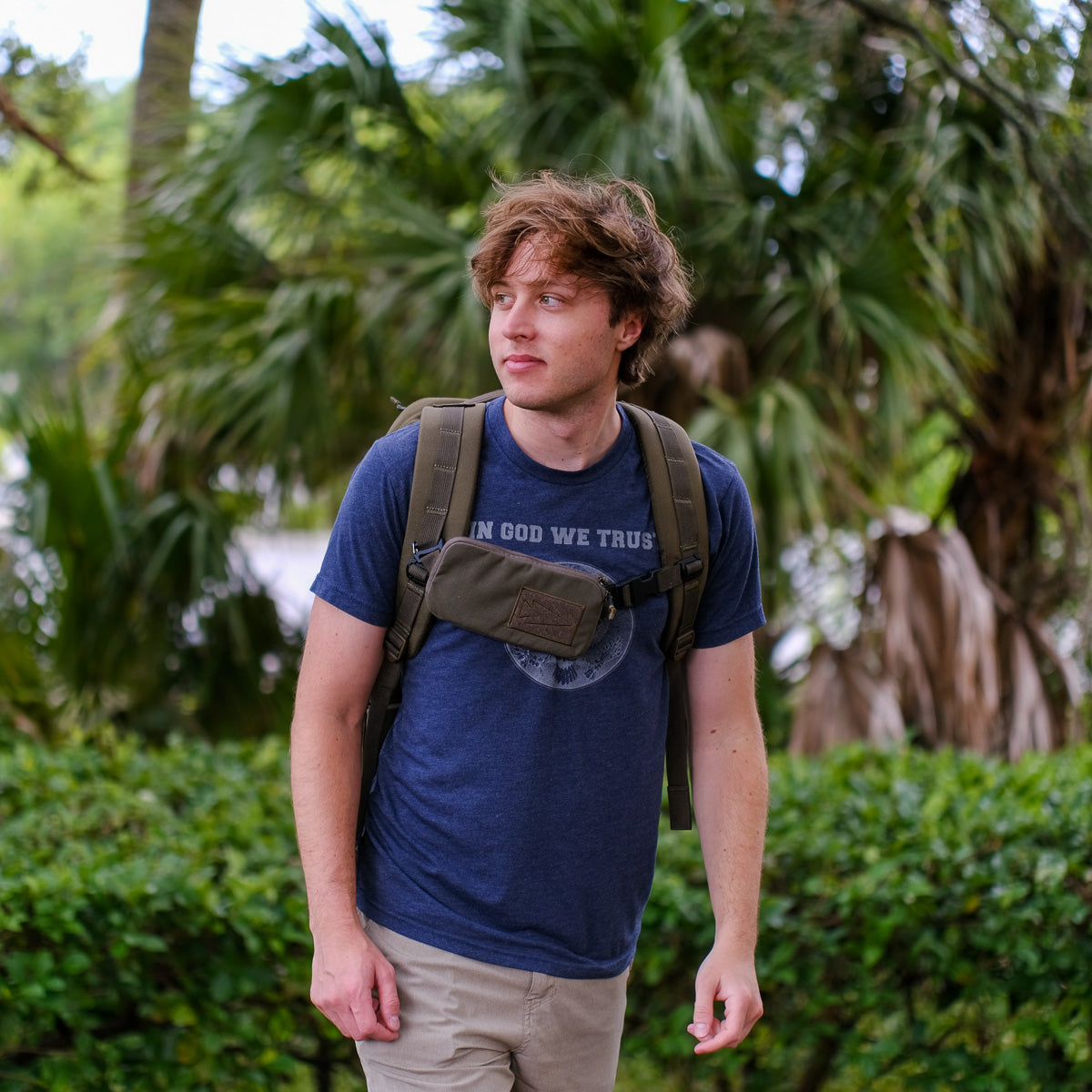 A young man with a backpack and a Sternum Pouch - Cordura stands outdoors among palm trees and greenery, looking to the side.