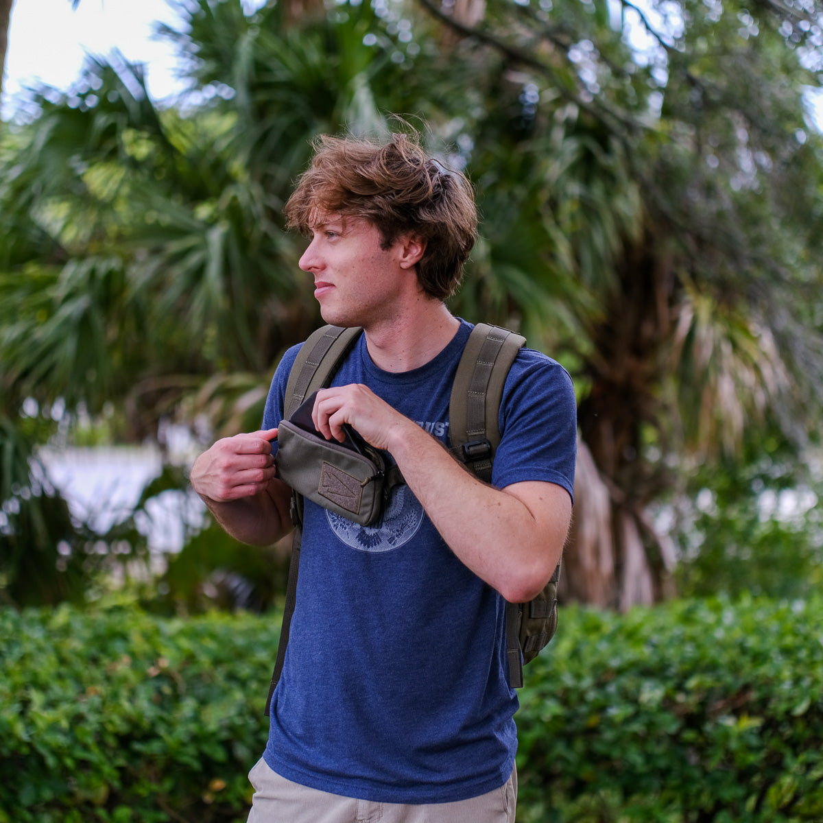 A young man with a backpack opens the Sternum Pouch - Cordura, a compact MOLLE-compatible pouch, while standing outdoors near green bushes and palm trees.