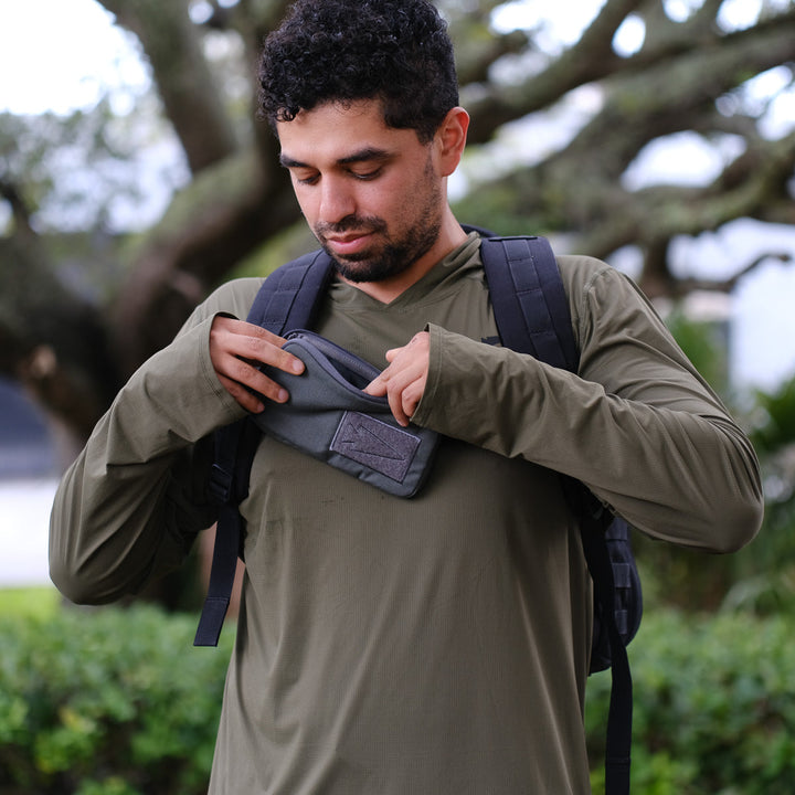 A man in an olive green shirt and backpack places a black wallet into the Sternum Pouch - Cordura, a MOLLE compatible pouch, while outdoors.