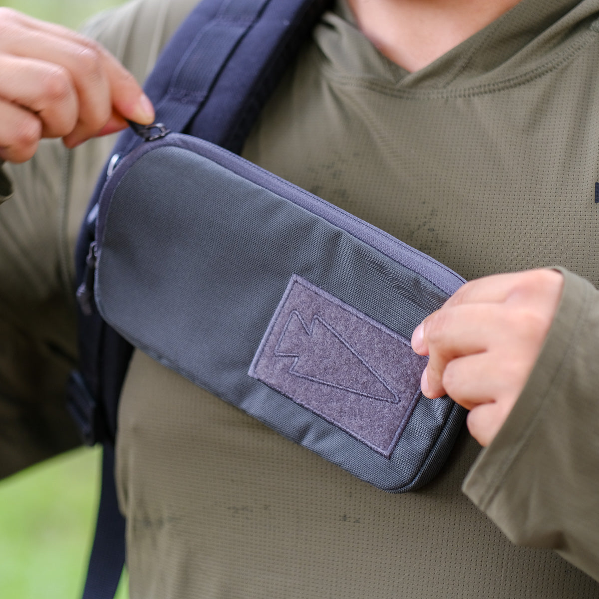 A person in an olive green shirt opens the Sternum Pouch - Cordura, a compact, MOLLE-compatible pouch attached to a backpack strap—an essential rucking accessory for added versatility.