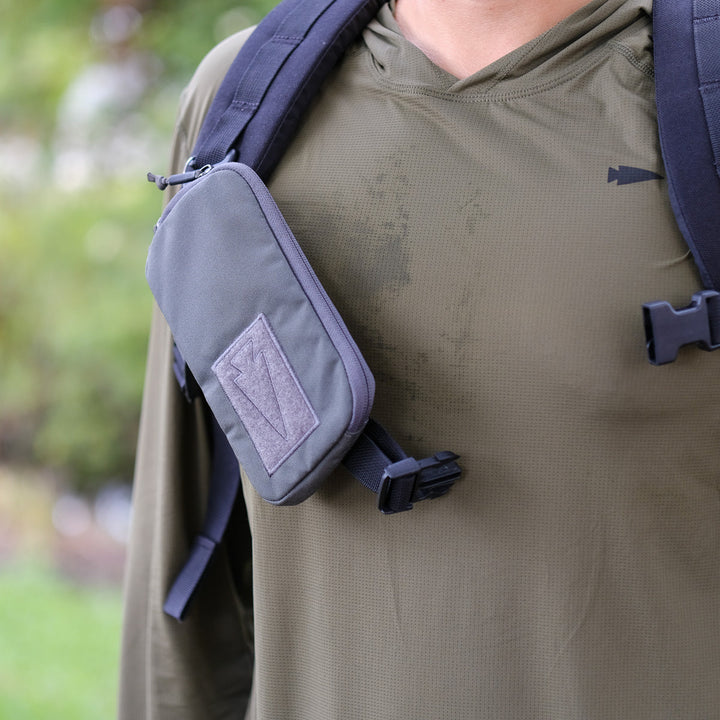 A person wearing an olive green hoodie and backpack with the Sternum Pouch - Cordura, a MOLLE-compatible rucking accessory, attached to the strap.