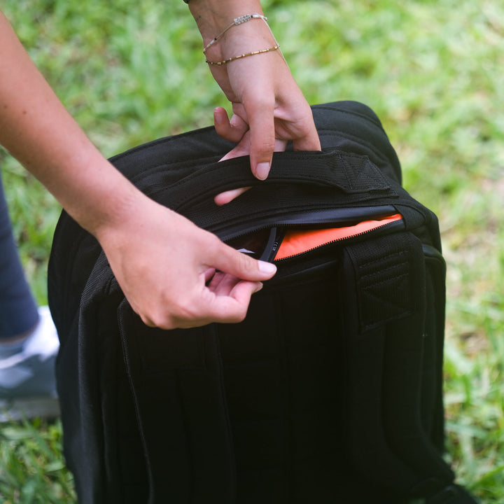 A person unzips the GR1 USA Slick - Ballistic Nylon Cordura backpack outdoors on grass, showing its orange interior pocket and bombproof laptop compartment—demonstrating quality American manufacturing.