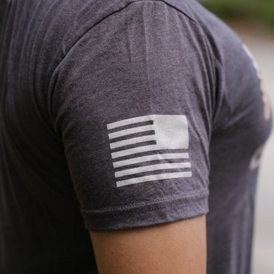 A white American flag graphic accents the sleeve of the GORUCK Logo DCU Camo Tee - Tri-Blend, a dark gray Special Forces-inspired T-shirt.