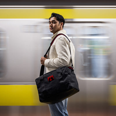 A man with a black Kit Bag x Carryology - GRXC2 Samurai 2.0 stands in front of a moving yellow subway train.