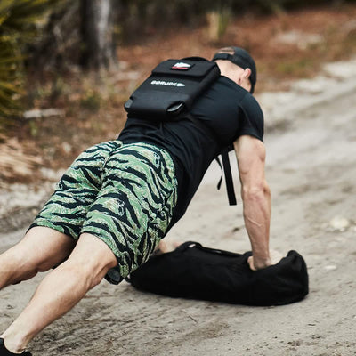A person wearing a black cap, black shirt, and green camouflage shorts is participating in a GORUCK event, doing push-ups on a dirt path. They have the Ruck Clubs Starter Kit - Heavy bag on their back and are supported by another bag in front as they ruck through the forest. Trees can be seen in the background.