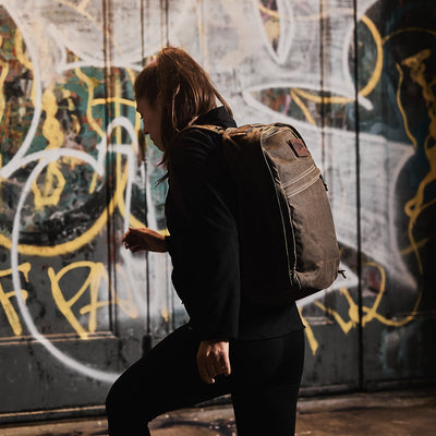 Someone carrying the GR1 USA - Heritage Waxed Canvas walks up steps in front of a graffiti-covered wall.