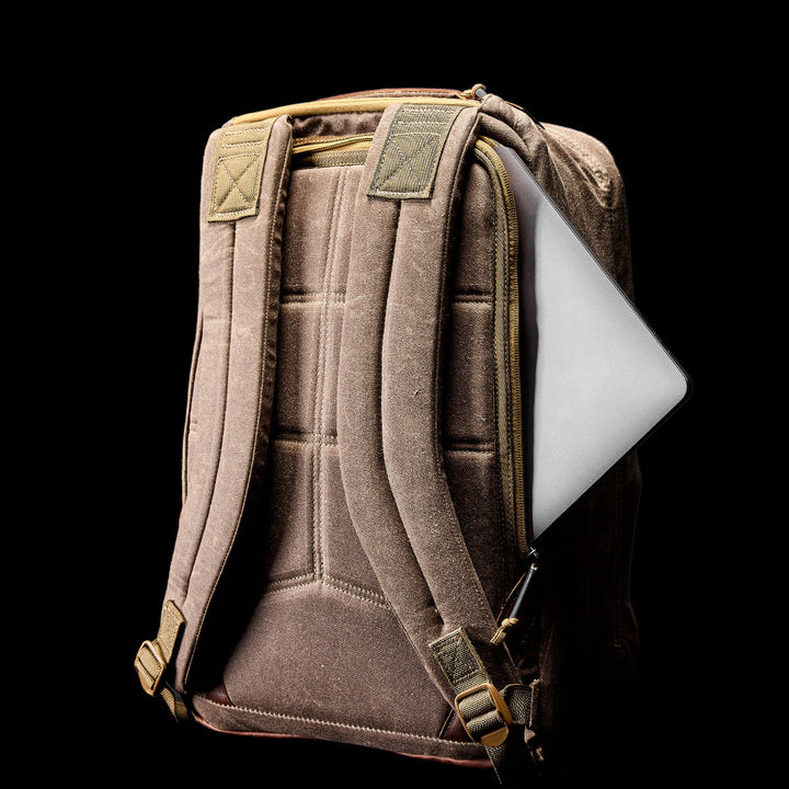 The GR1 USA - Heritage Waxed Canvas backpack in beige, with padded straps and a silver laptop partly visible in the side compartment, stands out against a black background.